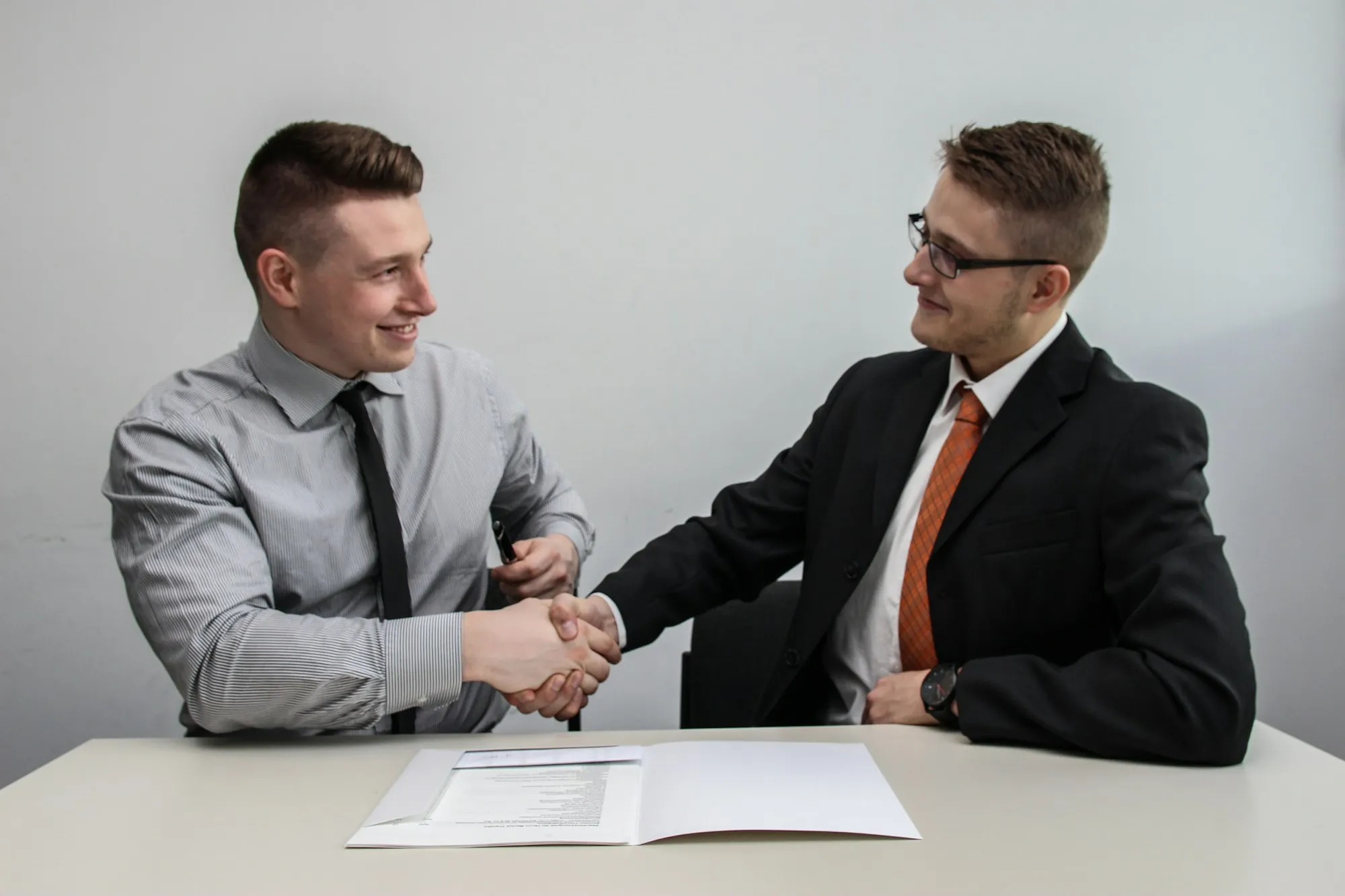 Two men in business attire shaking hands across a desk with paperwork
